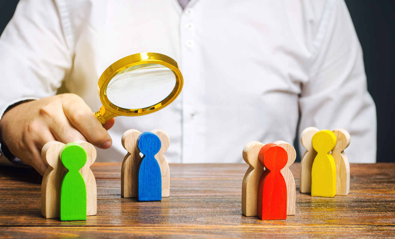 Person holding a magnifying glass over colorful wooden figures, symbolizing careful selection of beneficiaries for estate planning in Michigan.