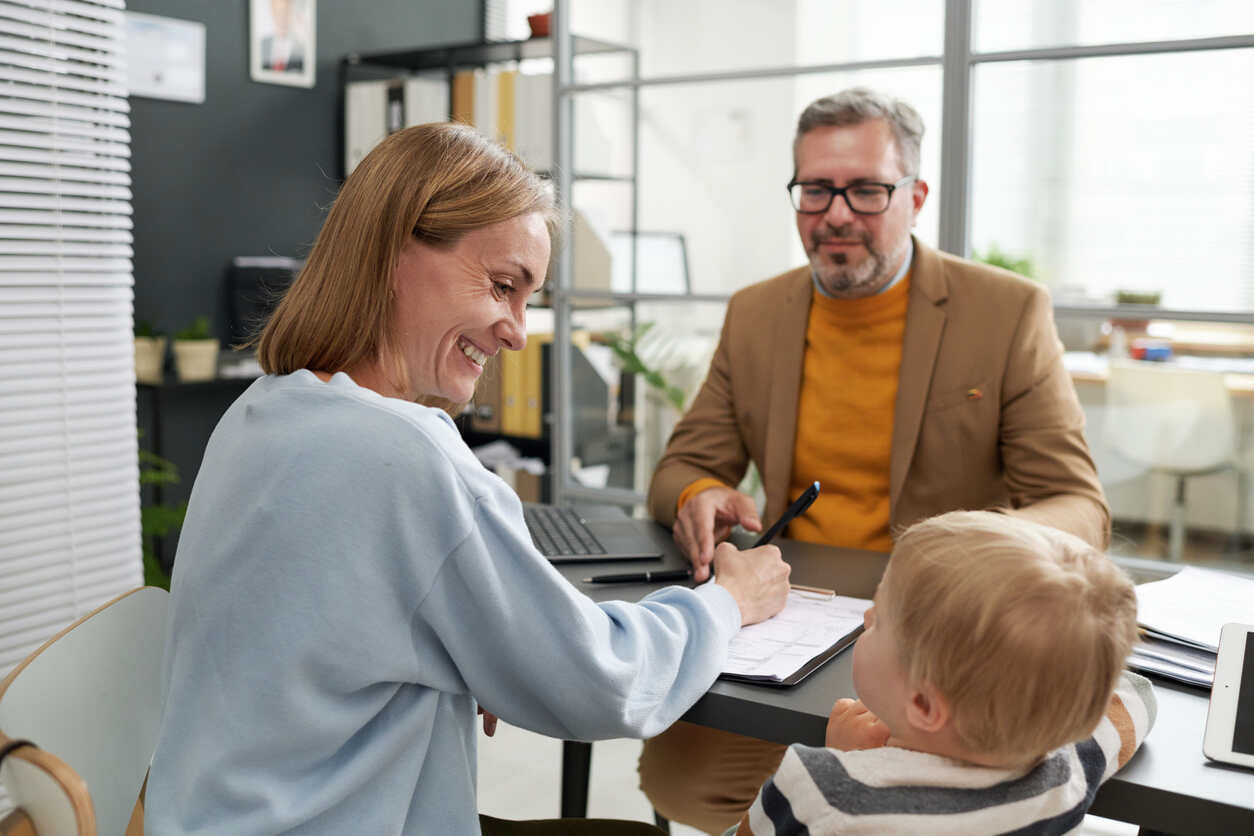 A smiling woman signing documents at a table with a lawyer and a baby present, symbolizing emergency guardianship planning.